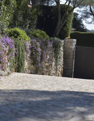 Paved driveway bordered by a dry-stone wall covered with trailing plants and purple flowers