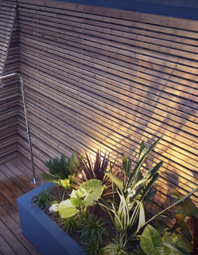 Terrasse en bois avec mur à claire-voie, douche extérieure et jardinière éclairée près d’une piscine