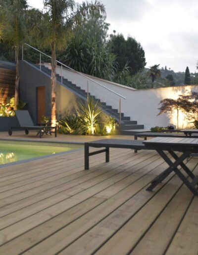 Terrasse en bois avec piscine éclairée, transats et plantations lumineuses en soirée