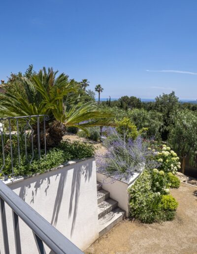Terrasse extérieure surélevée avec palmiers, massifs fleuris et vue dégagée vers la mer